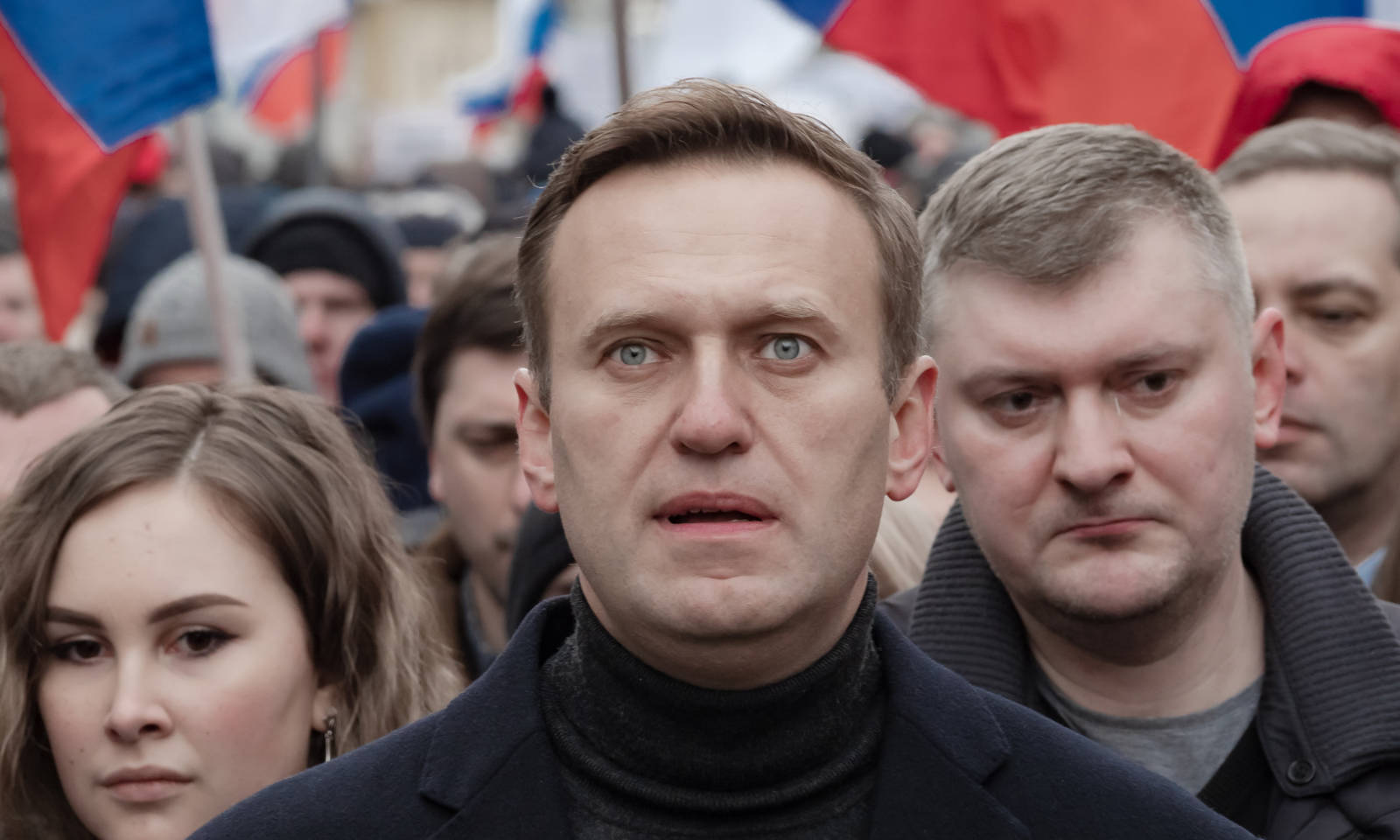 Alexei Navalny staring ahead with a crowd of people behind him holding Russian flags.