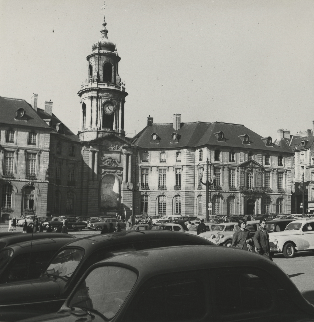 black and white photo of two buildings next to each other with a taller clock tower between them and a parking lot of cars in front of them all