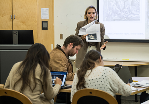 A professor explainging a slide on a projector screen to a class.