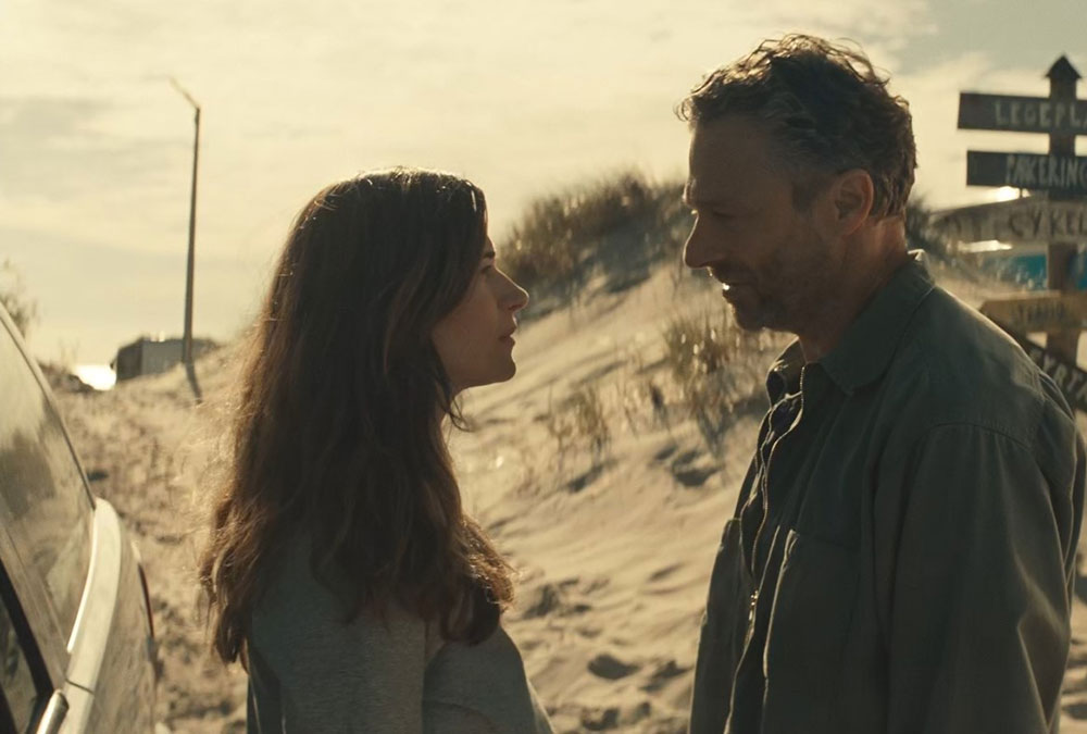 A still image from the movie of a man and woman facing each other while standing next to a car on the side of the road.