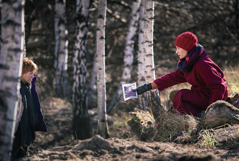 A still image from the movie of a woman wearing a red hat and coat handing a photo to a young boy in the woods.