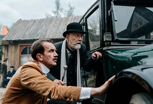 A still image from the movie of two men leaning against an older car.