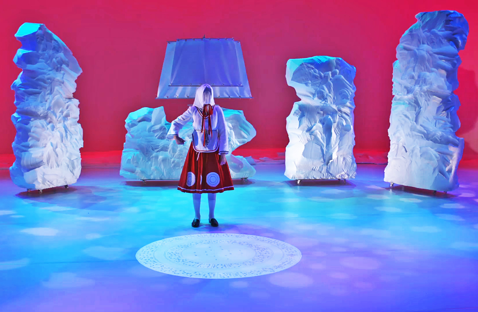 A still image depicting a person on stage in costume with their face covered.
