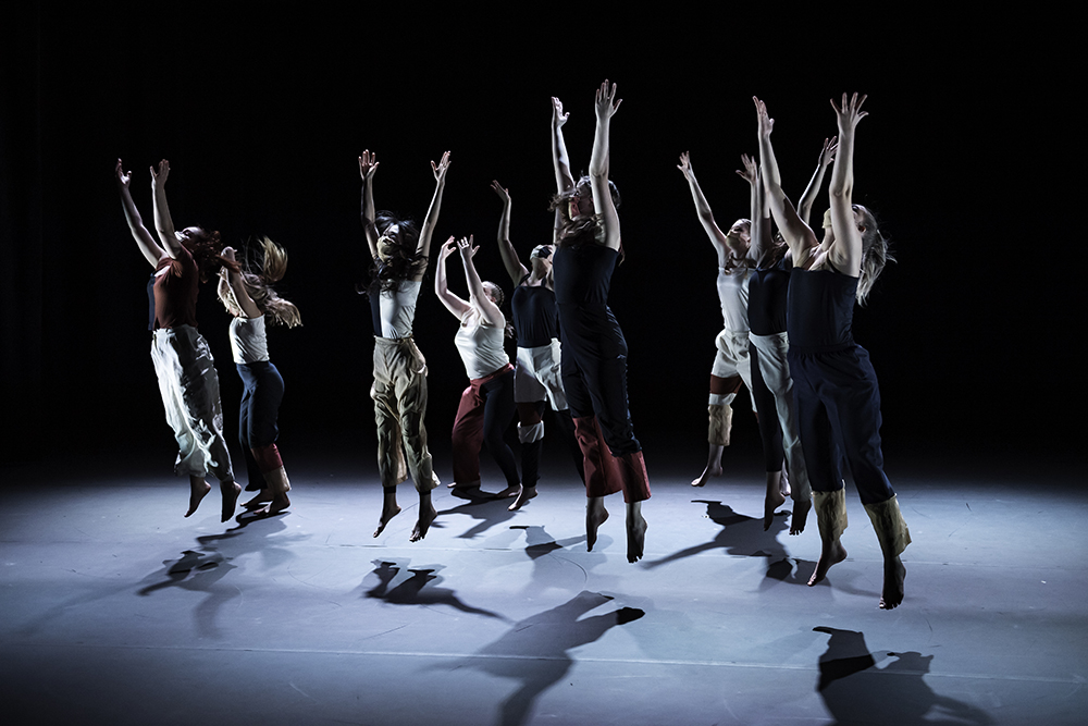 A group of dancers reach above their heads on a darkened stage.