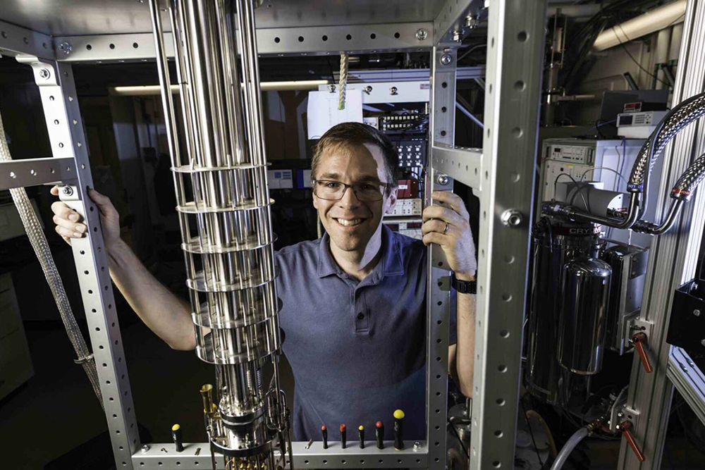 John Nichol standing behind a dilution refrigerator, used to study quantum systems, including nuclear-spin dark states.