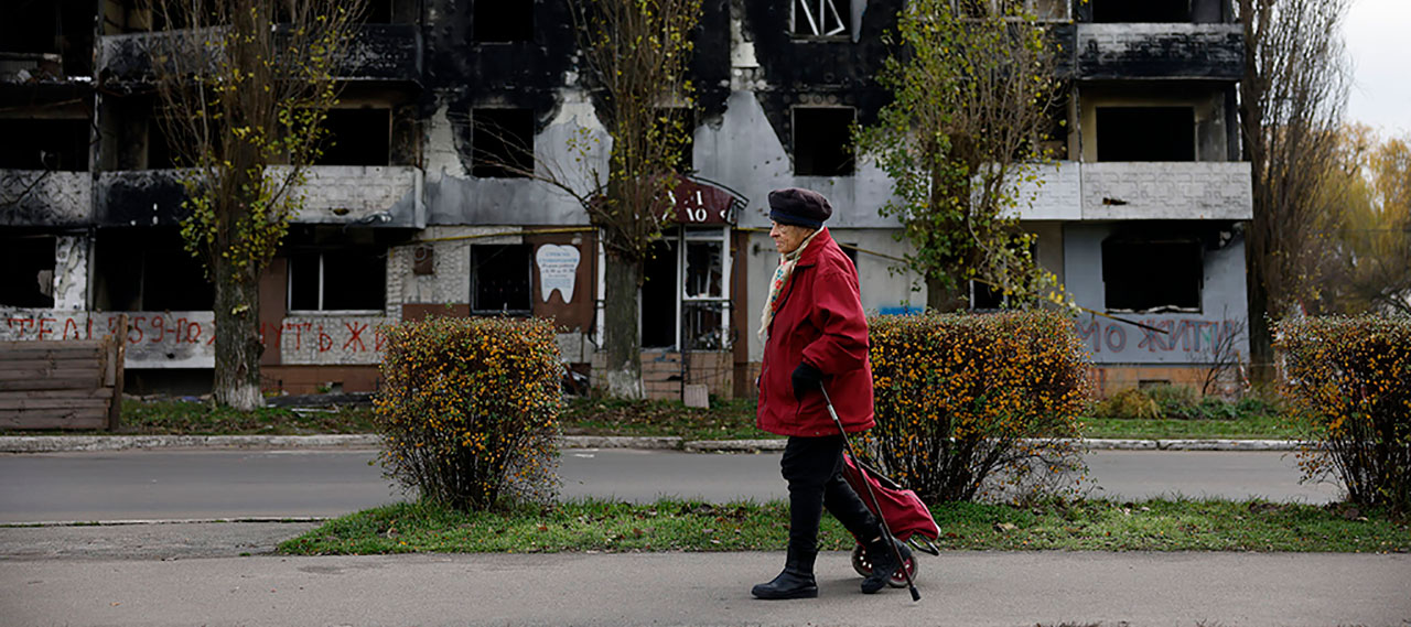 A Ukrainian in Borodyanka goes about daily life walking down a sidewalk with a bombed out building in the background.