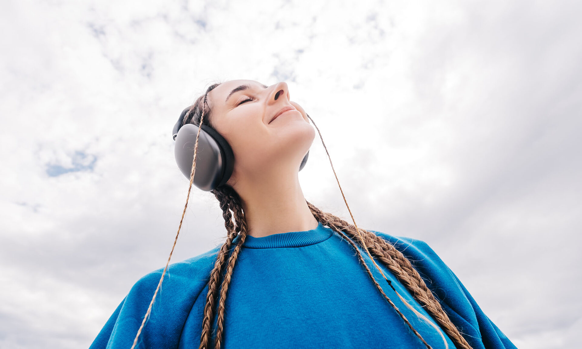 A young woman with pigtails wearing headphones and a blue sweatshirt is listening to music against the sky from a low wide angle.