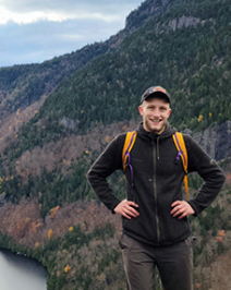 William is posing for the camera outdoors with a mountain range in the background.