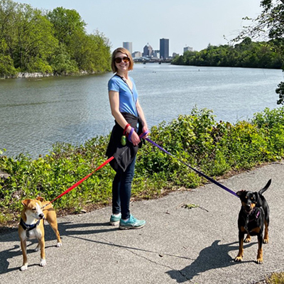 Rose smiling at the camera while holding two leashed dogs with the Rochester skyline in the background.