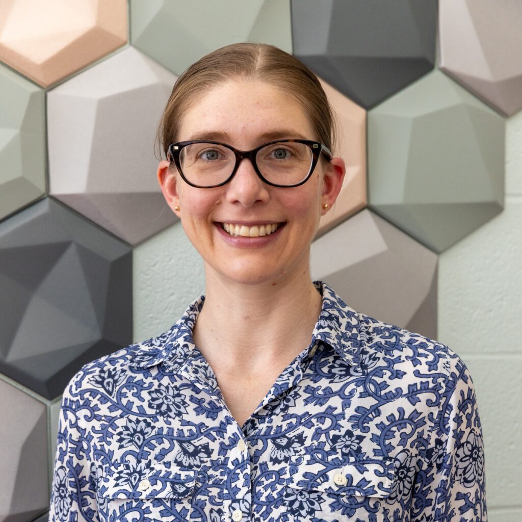 A woman with glasses and a patterned shirt standing in front of a wall of hexagonal tiles