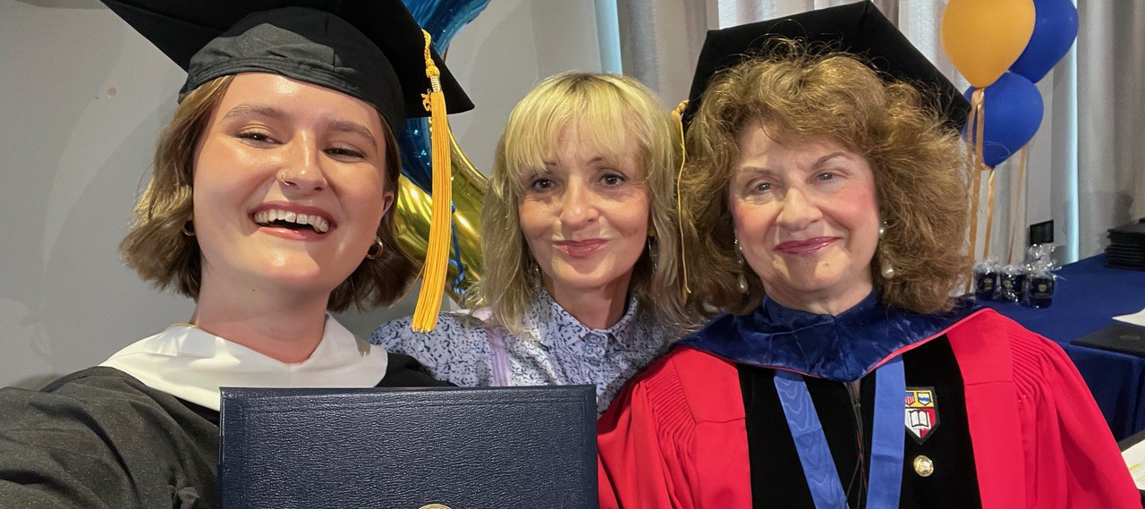 A student in graduation attire holds a diploma up to the camera white two other people look on.