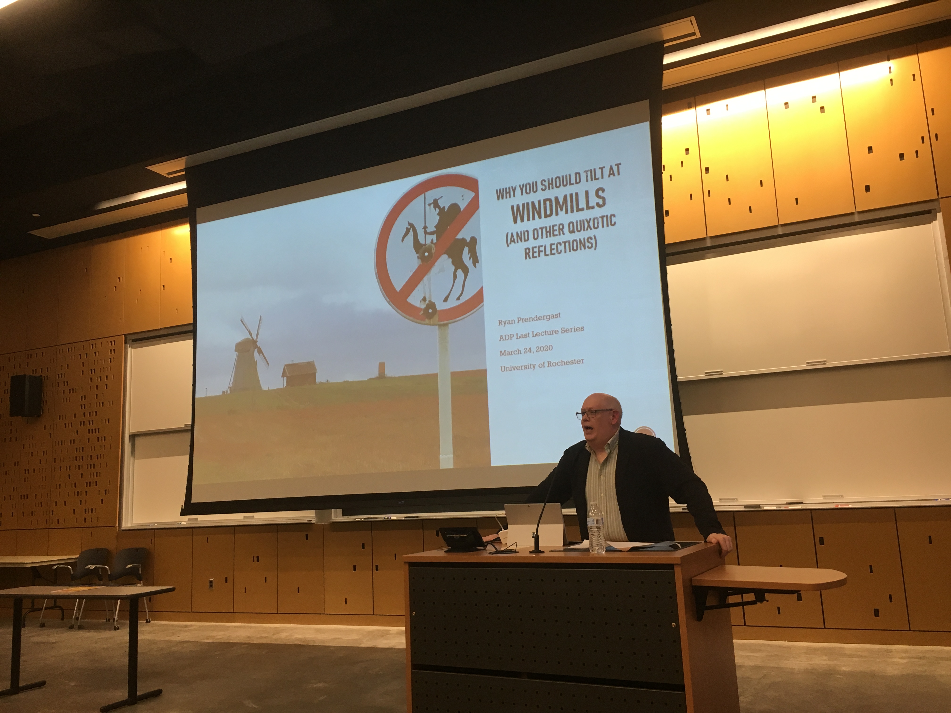 Dr. Ryan Prendergast stands at the podium in front of a projected powerpoint presentation at his lecture