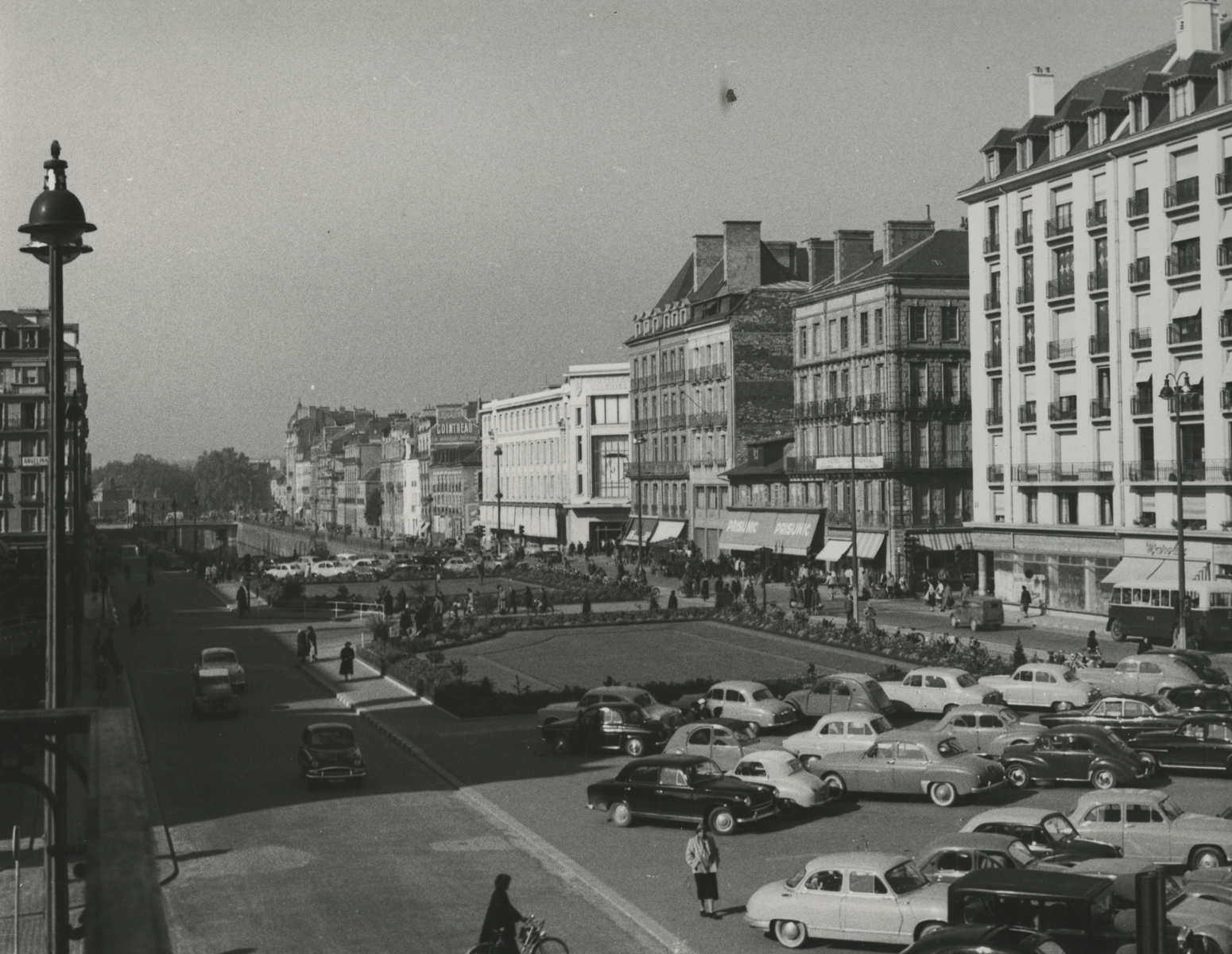 black and white photo of buildings stretch into distance on right, various cars parked near a grassy area between two parallel roads