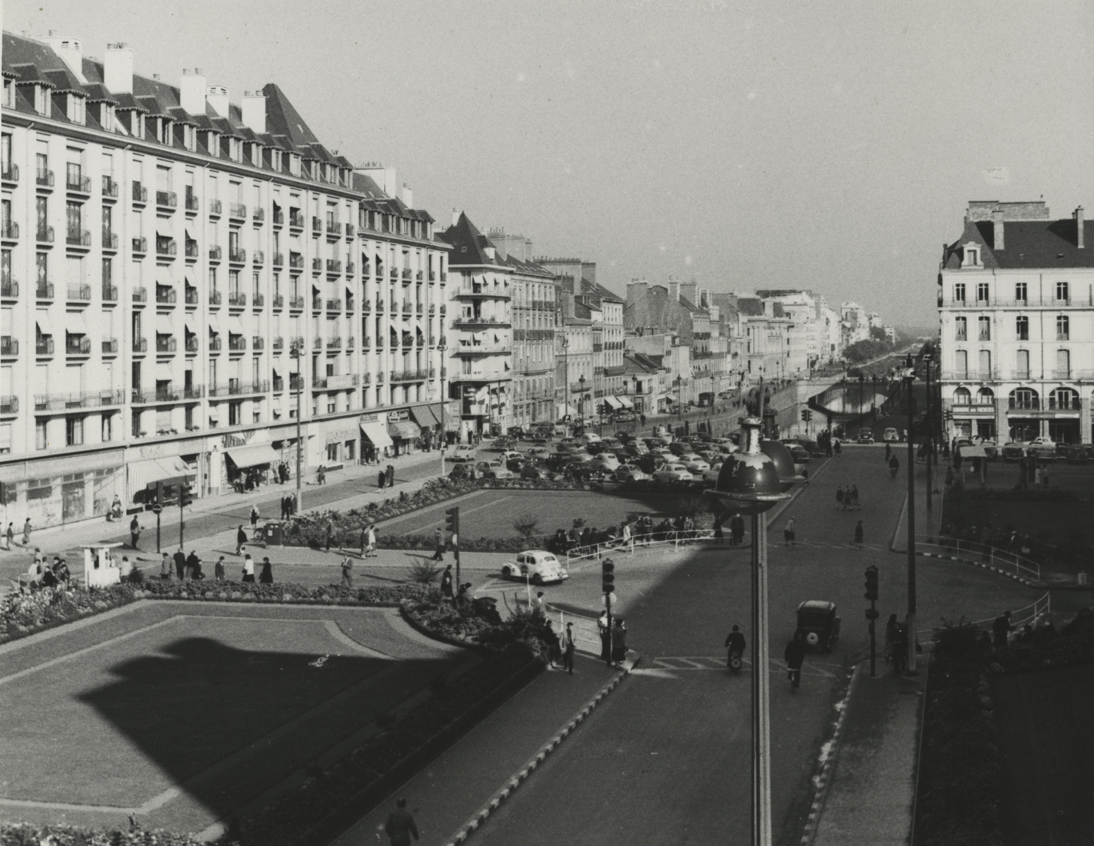 black and white photo of buildings stretch into the distance on the left in front of parallel roads with grassy areas and parked cars between them