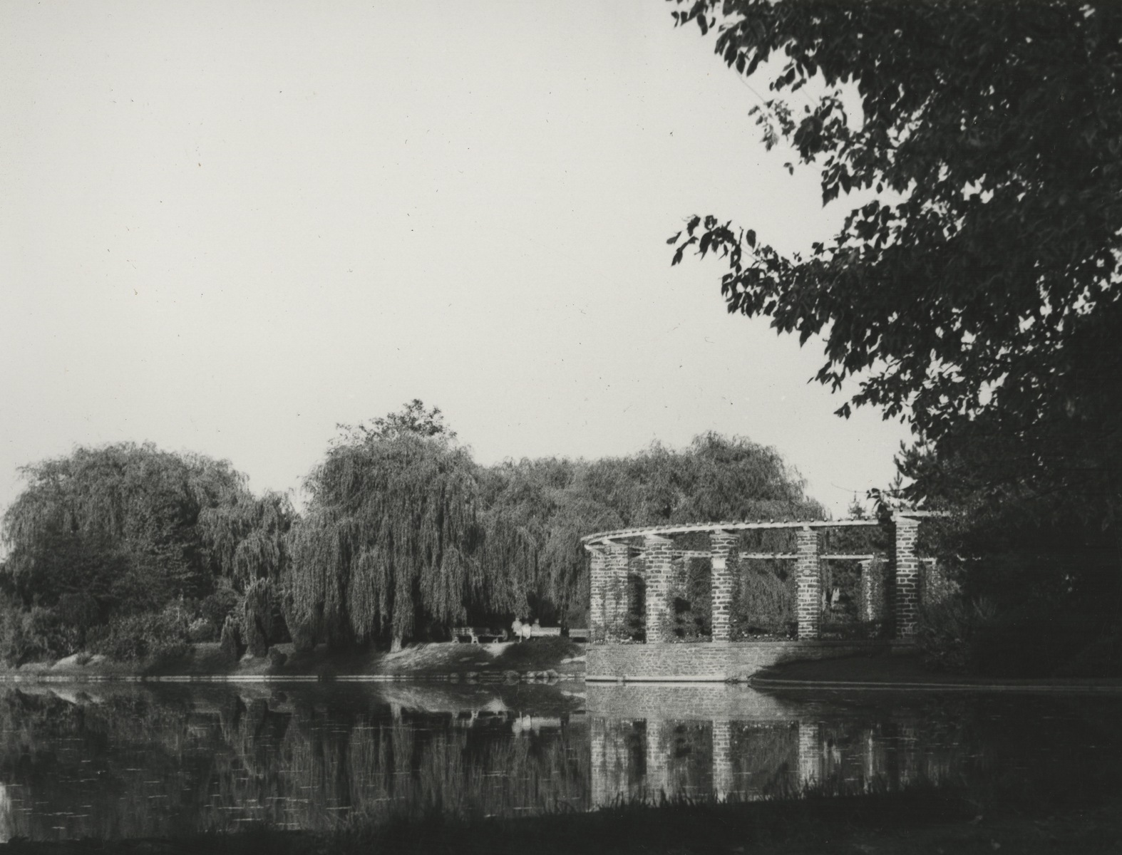 black and white photo of a brick gazebo in a large pond with willow trees along the far bank