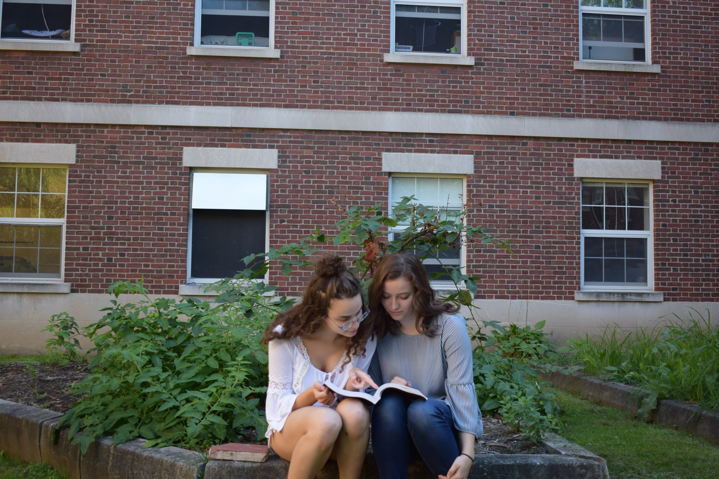 two girls share a book while sitting in a garden behind a brick building