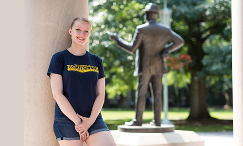 Lucy Farnham stands leaning against a pillar with her back to the Eastman Quadrangle