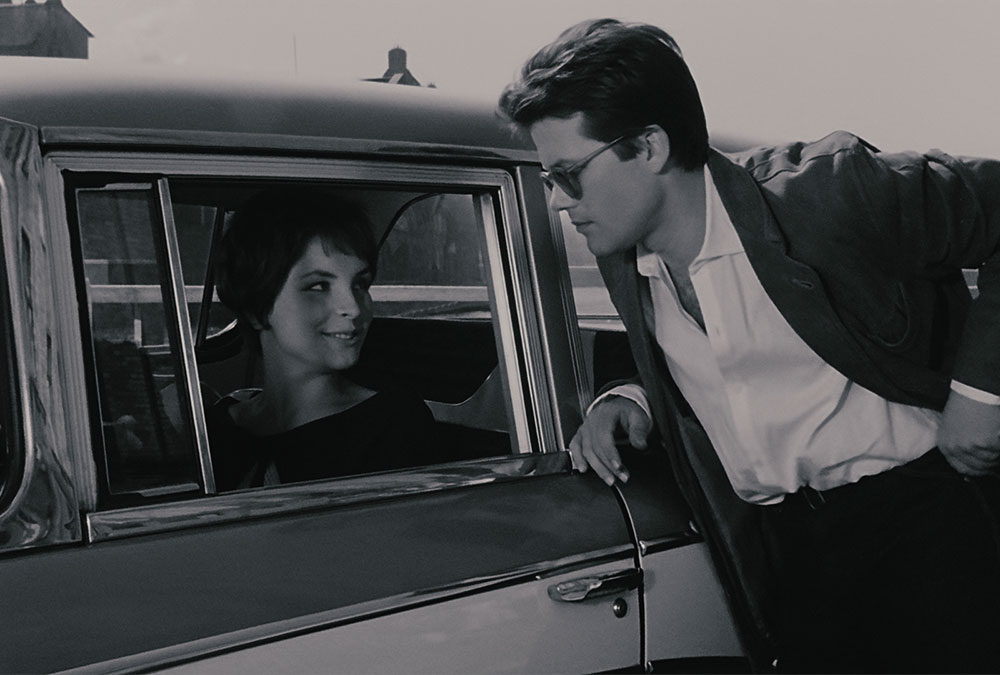 A still image from the film of a man leaning against an open car window speaking to a woman inside the car.