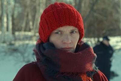 A still image from the film of a woman wearing a red hat staring at the camera with a person in the background behind her.