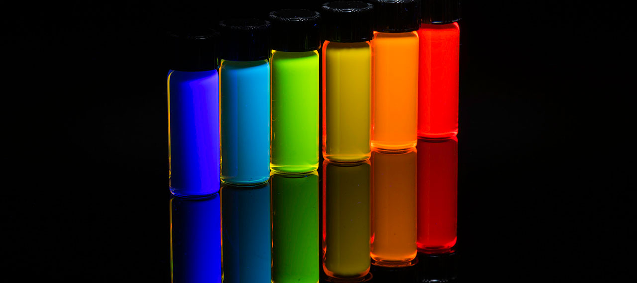 Six vials of quantum dots fluoresce in bright colors against a black background.