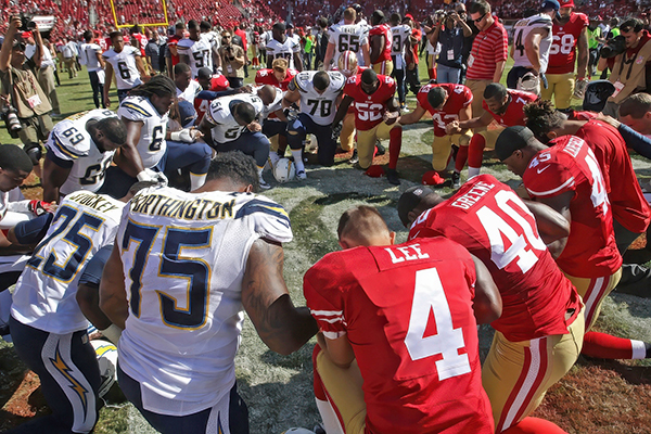 Football players in uniform kneeling together in a circle on the field with their heads bent in prayer.