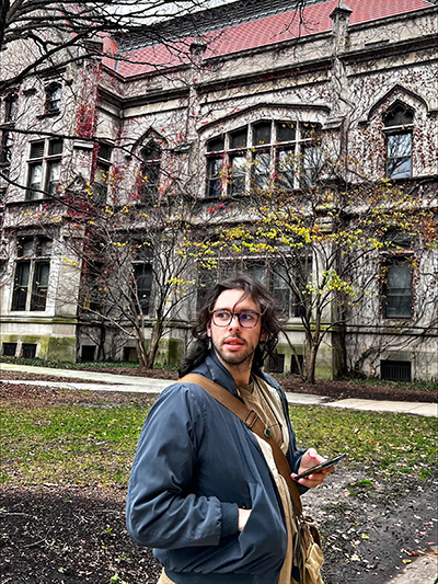 Liam Kilby looking over his shoulder while holding a cell phone with a large building in the background.